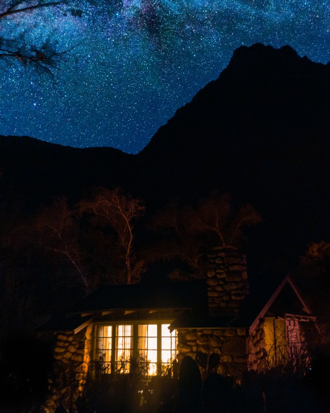 A small cabin built primarily of field stone sits at the bottom of the frame, barely visible in the dark. Yellow light streams from two window openings, which have some curtains behind their multiple panes. You can make out the leaves of a tree stretching above the cabin into the dark silhouette of a tall craggy mountain or ridge. The sky is filled with white, green, and purple stars that get more dense toward the top of the frame. 