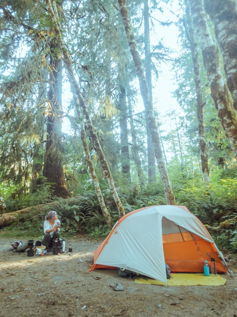 Laura sits on a rock surrounded by our backcamping food supplies. Our orange and grey Big Agnes tent is set up just next to her. The trees of the Hoh Rainforest tower above. 