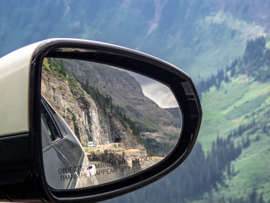 Most of the frame is taken up by a car's side mirror, which shows a reflection of a sheer rock face, some cars driving along a road at its base, and a tunnel into the rock in the distance. Visible around the edges of the mirror is an out-of-focus landscape in the distance, which appears to be a steep mountainside covered in green grass and spruce trees. 