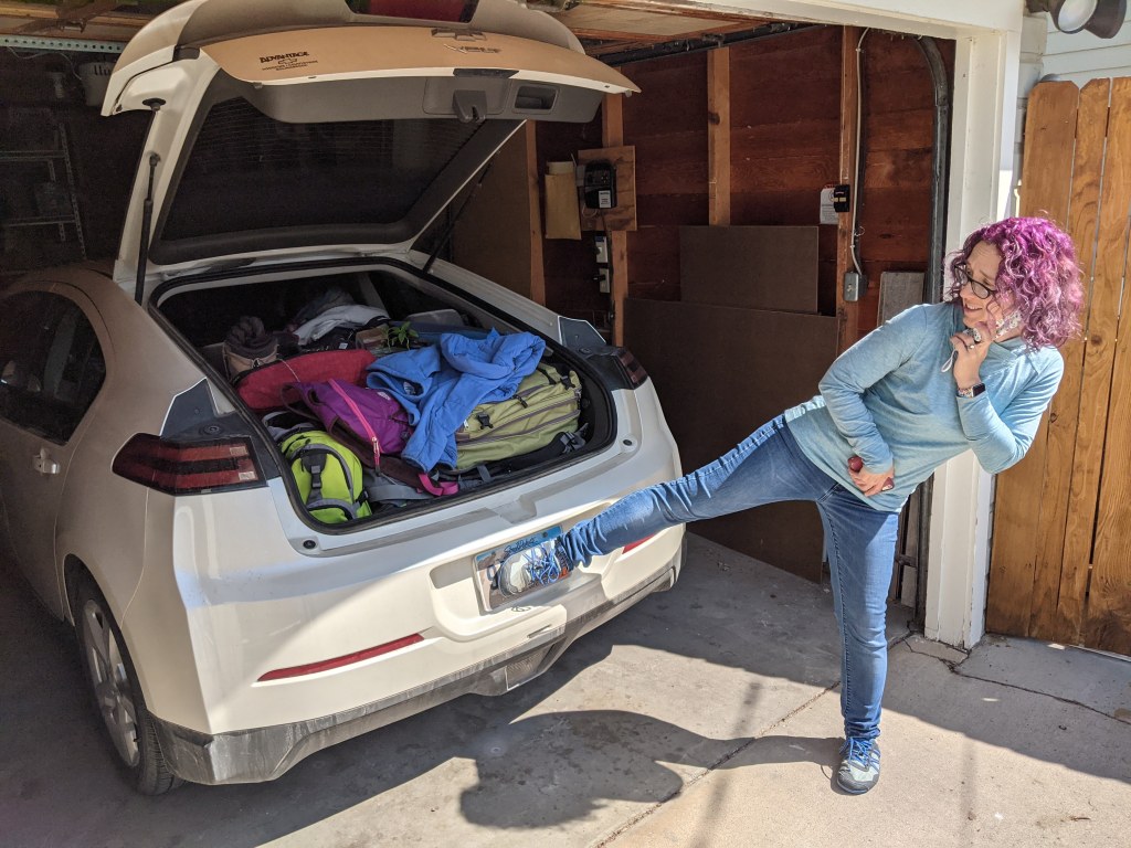 Our white Chevy Volt is parked in our garage with the hatch left open. It's pretty obviously packed full, with various backpacks, coats, and miscellaneous objects arranged Tetris-style to fit in the available space. Laura is at the right side of the frame, holding her right foot over the license plate to obscure the number for the sake of the photo. 