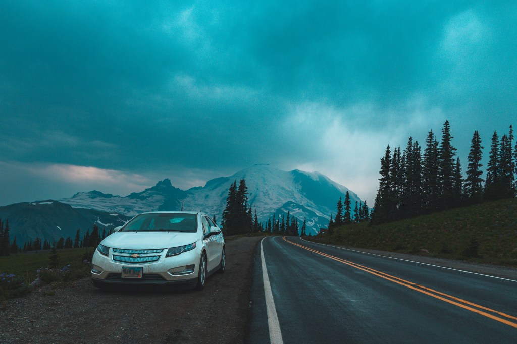 In the left half of the frame, our white 2013 Chevy Volt is parked on the side of a road that curves off into a stand of fir and spruce trees on the right. In the background, you can see the snow and glacier-covered sides of Mount Rainer. Clouds with a blue and pink tone hang around the slopes, but you can see the summit. 