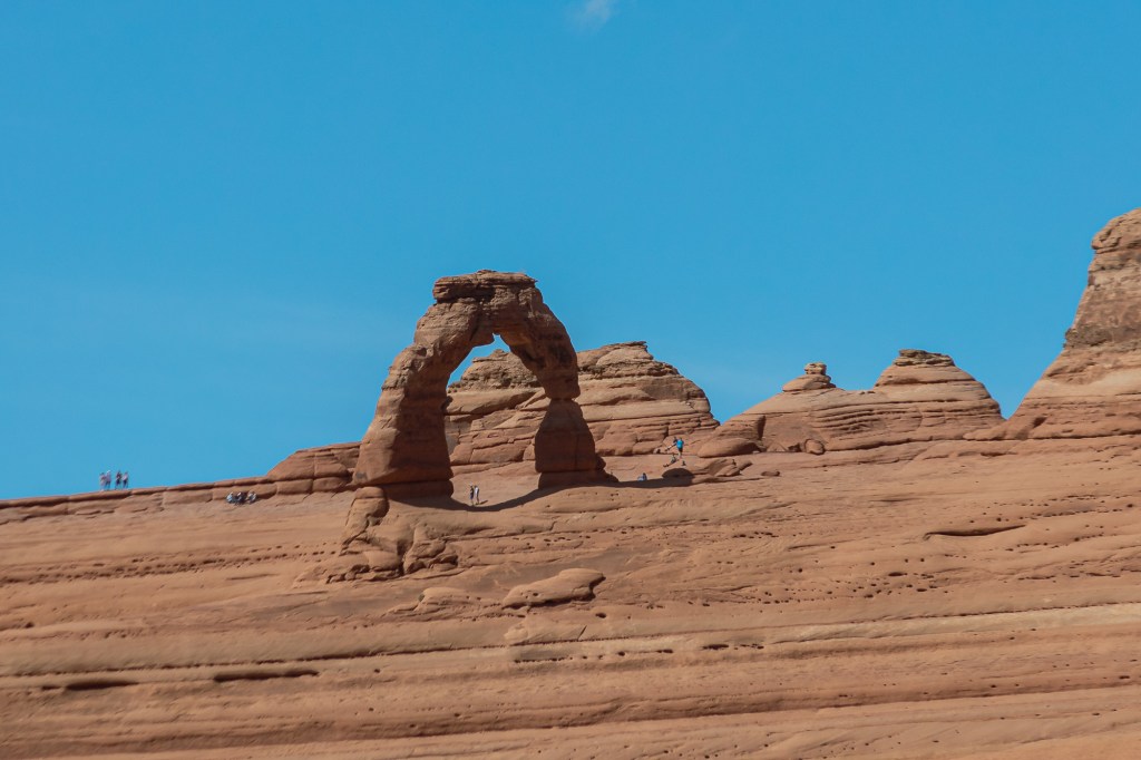 A telephoto view of Delicate Arch, which stands at the center of the frame surrounded by a small crowd of hikers. Some sandstone ridges and formations fill out the rest of the image, with a clear blue sky above. 