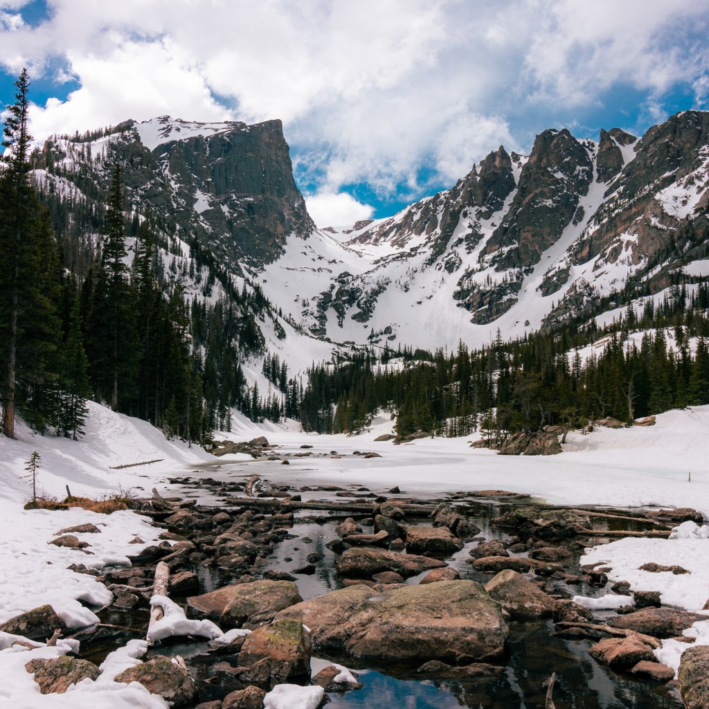 At the bottom of the frame, some rocks and water are visible against an otherwise snow-covered landscape that stretches into some stands of spruce and fir trees in the middle-distance. Sharp rocky mountains suddenly rise up in the background, touching a sky filled with fluffy white clouds and only a few patches of bright blue. 