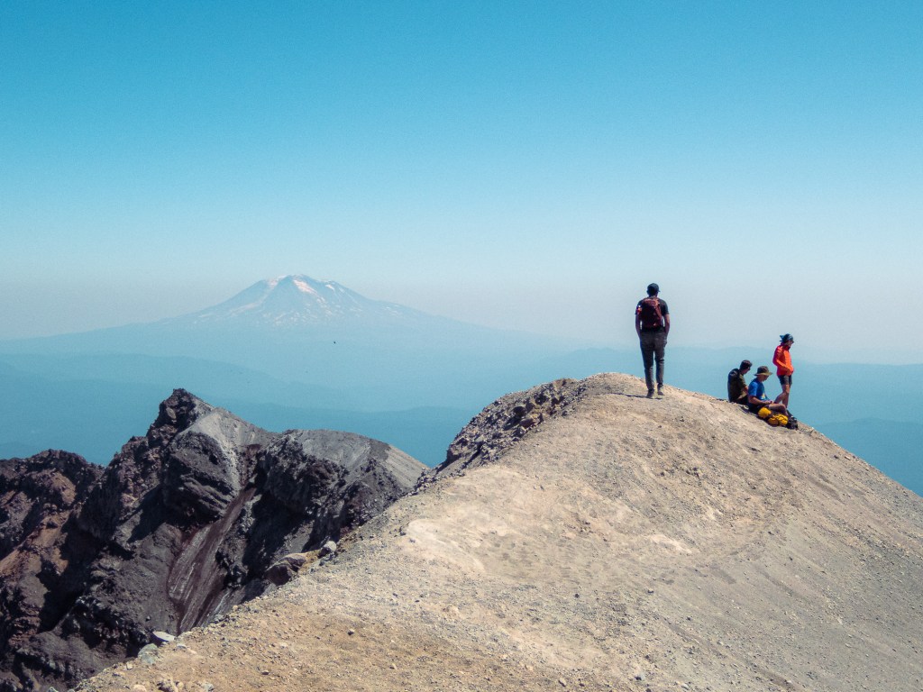 In the middle of the frame, a group of hikers congregate on a narrow ridge of yellow rock on the top of Mount Saint Helens. In the distance, through some haze, the peak of Mount Adams is visible, towering over the landscape against a bright blue sky. 