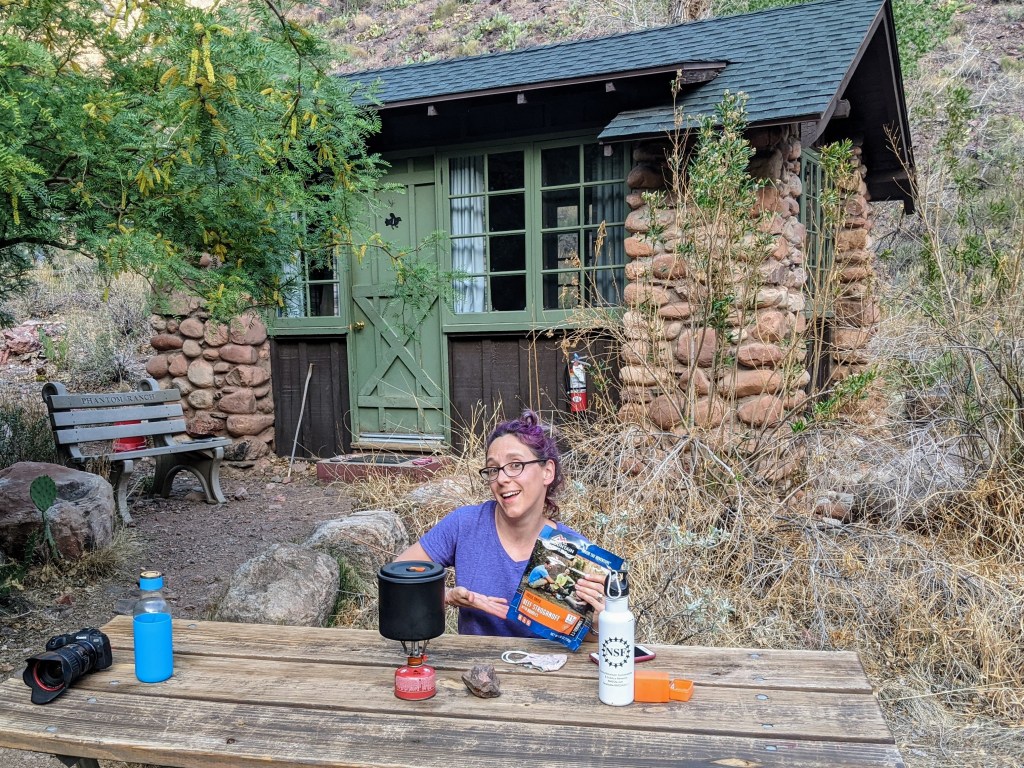 Laura sits at a picnic table at the bottom of the frame, gesturing toward an envelope of freeze-dried beef stroganoff. There's a pot sitting on top of a gas burner nearby, along with some water bottles and my black Canon camera. In the background, our cabin takes up the rest of the frame. It's got large fieldstone pillars on the corners, with brown wood siding in between. There are large green-trimmed multi-paned windows the entire distance between the front pillars, broken only by a green cottage-style door. 