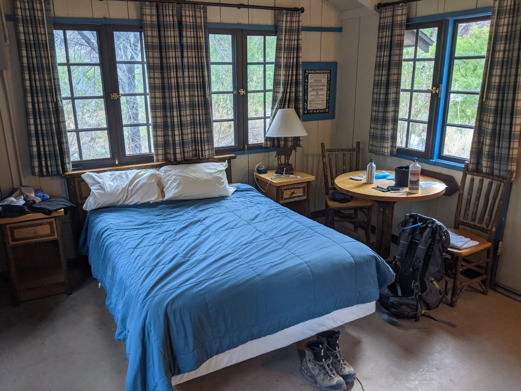 A corner view of our cabin. A queen bed fills most of this part of the room, covered in a simple blue blanket. There are two rustic bedside tables, a cast-iron lamp in the shape of a horse, and a small table and two chairs nearby. There are three window openings above the bed, and each one has two french-style windows divided into eight small panes. There are brown grasses and green trees visible outside.  A pair of boots and a backpack rest on the floor, and there are some water bottles and a mess kit on the table. 