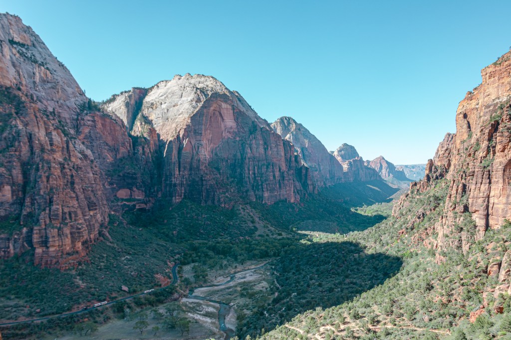 The landscape of Zion Valley stretches off into the distance, with the rock temples standing sharply against a clear blue sky. The valley bottom is mostly cast in shadow, but it's possible to see the Virgin River and park road - with two shuttles - winding their way between the mountains. 