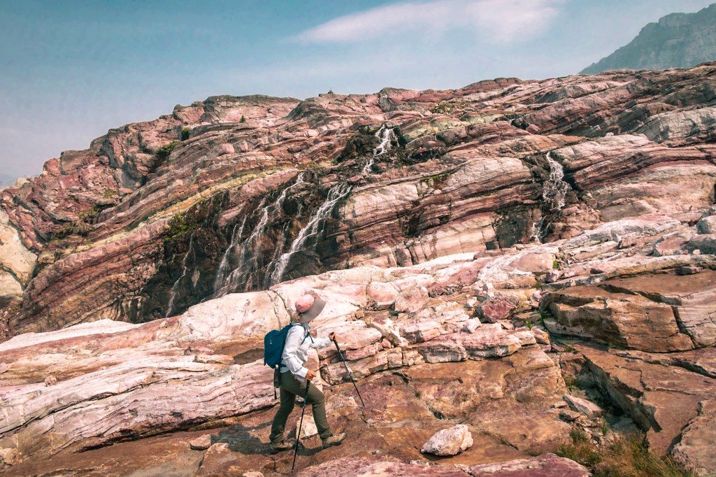 Rocky ledges striped white and yellow and worn smooth by glacial erosion fill the background. A small waterfall makes its way down the center of the frame toward Laura, who stands at the bottom looking toward the waterfall.