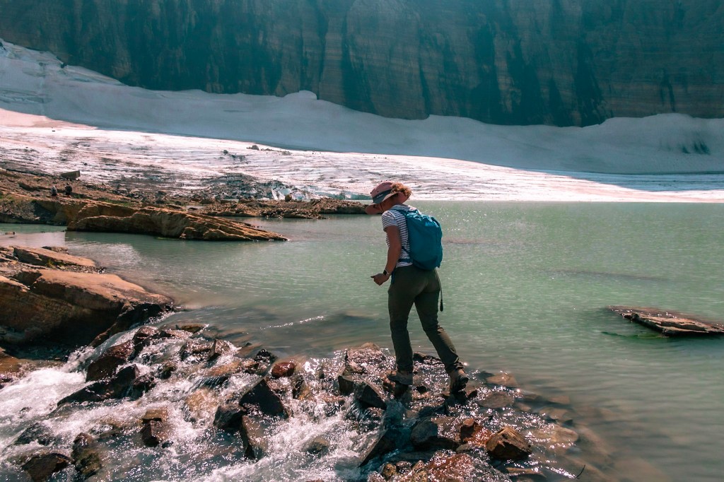 Laura is in the center of the frame, walking over some rocks that have water cascading over them. A lake sits just behind the cascade, with the bright white snow and ice of a glacier visible in the background. 