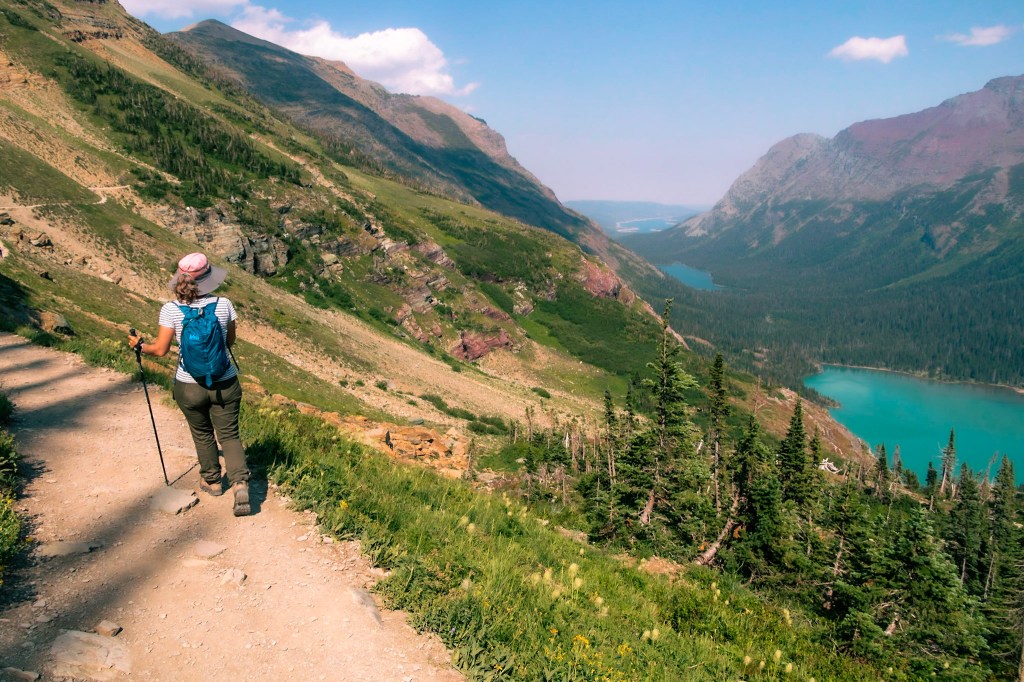 Laura walks along a rocky trail at the left of the image. To the right, there are three bright teal lakes visible along a valley floor, which stretches off into the distance. 