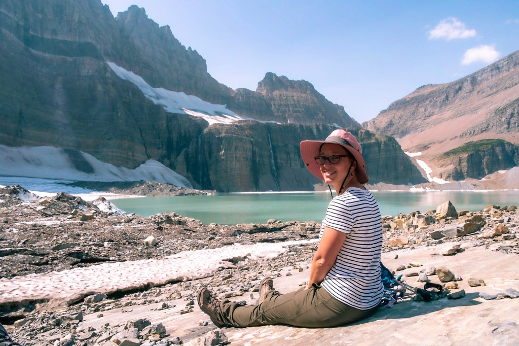 Laura sits on a bare rocky patch looking at the camera. The blue/green waters of the glacial lake are immediately behind her, with the ice-topped mountains rising above. 