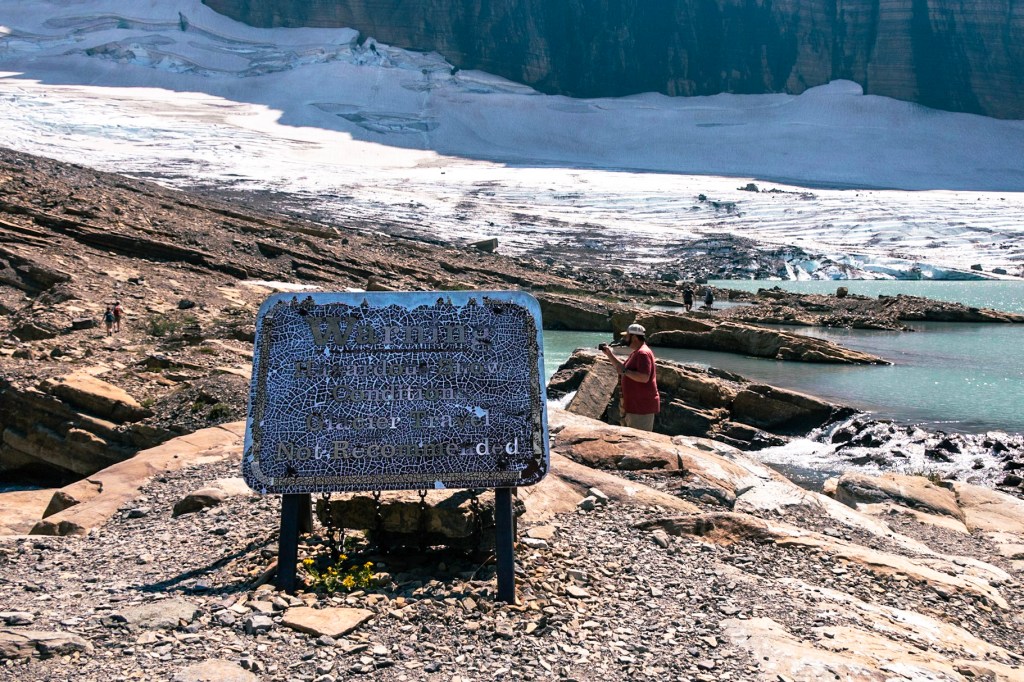 A badly worn sign sits on a pile of talus in the center of the image. The words, "Warning: Hazardous Snow Conditions, Glacier Travel Not Recommended" are just barely visible. In the background, you can make out some people along a rocky lakeshore, with a large glacier and rock wall immediately behind them. 