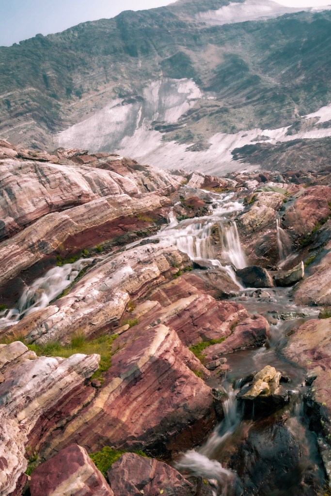 A vertical photo showing a landscape of grey, white, and red banded rocks being run through by a number of braided waterfalls. 
