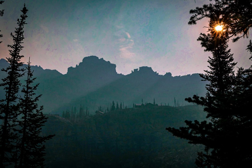 The sun, in the top right corner of the photo, shines orange through the branches of a spruce tree. Dark mountains in the background with a darker ridge closer to the foreground, upon which the silhouette of buildings (Sperry Chalet) can barely be discerned. 