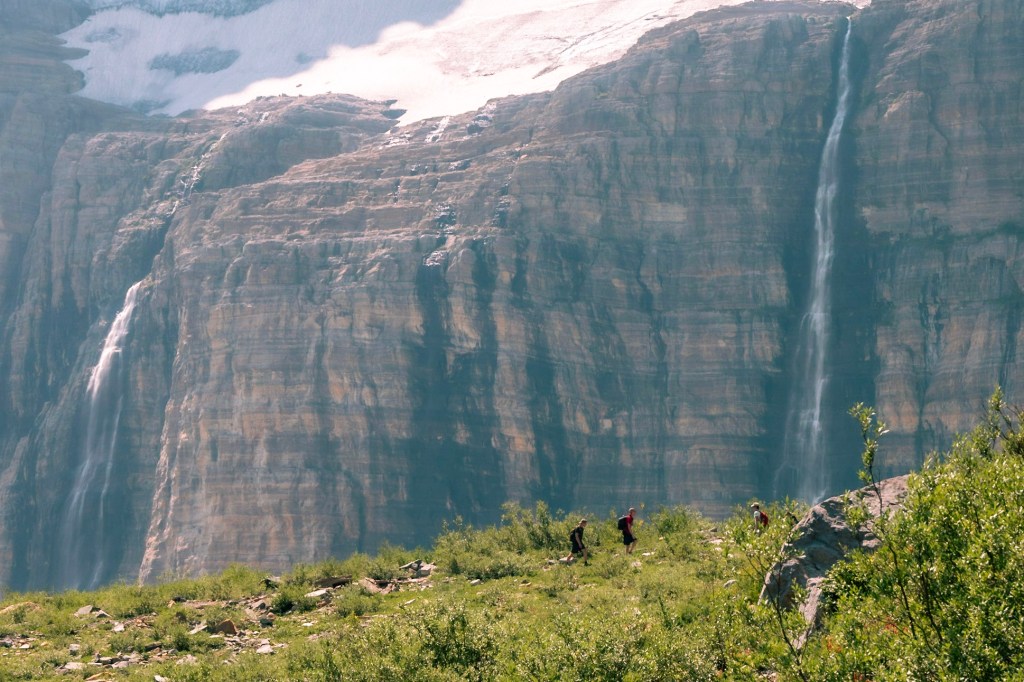 In the foreground, there are outlines of three hikers walking along a trail. The background, which dominates the image, is a sheer rock wall, with a mass of snow and ice sitting on a ledge at the top. Cascades of water stream down from the ice along the rock wall. 