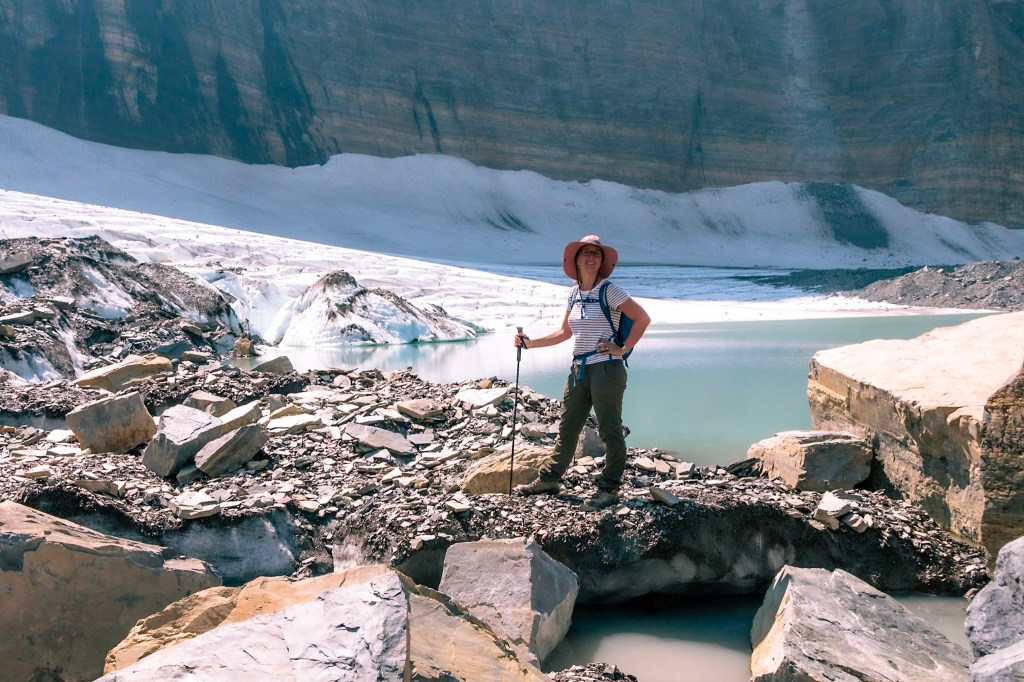 Laura stands in the center of the image, proudly perched on a pile of rock, ice, and snow. Grinnell Glacier and the glacier's lake stretch to fill the frame behind her. 