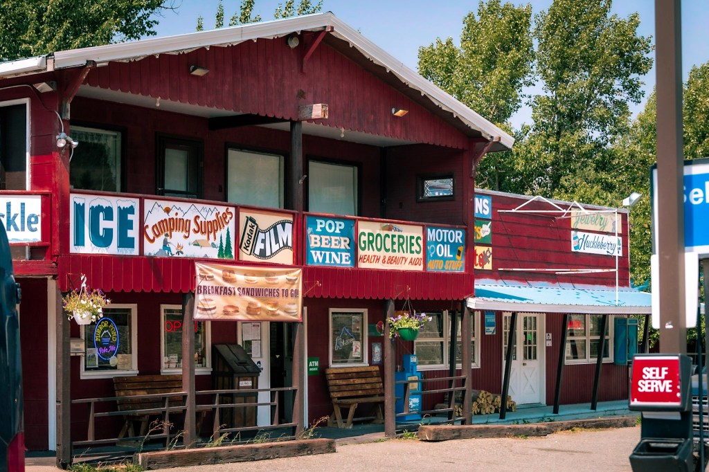 A building covered in red board siding fills the frame. It is two stories tall with a peaked roof. An entry door is at center on the lower level, framed by windows fill of neon signs. Above the door across the whole front of the building stretch signs for "ICE", "Camping Supplies", "Kodak FILM", "POP BEER WINE," "GROCERIES Health & Beauty Aids", "MOTOR OIL Auto Stuff" and "Jewelry", and "Huckleberry." 