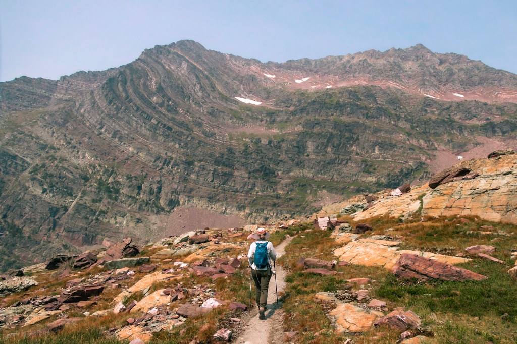 Laura hikes away from the camera at the bottom center of the frame. The path she follows winds between orange and red rocks toward a mountain with wildly folded sedimentary layers in the background.