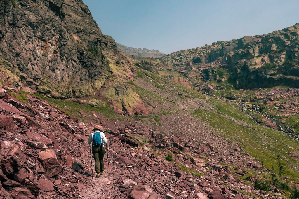 A stretch of treeless mountainside which slopes steeply up on the left. In the distance, another slope rises to the right. Laura walks along a stone path in the bottom left. 