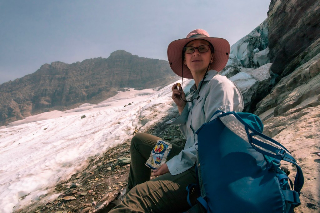 Laura sits off-center on gray rock, with Sperry Glacier visible on the left. She's happily munching Pirate Booty.