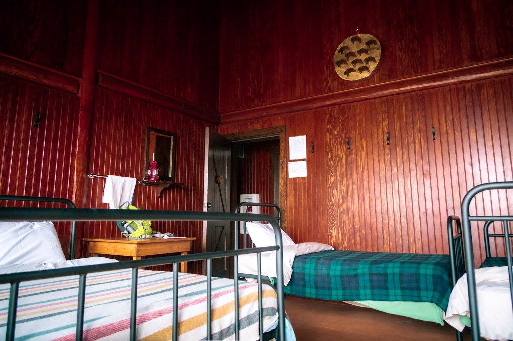 The interior of a dorm room at Sperry Chalet shows a double bed on the left and a single bed each against the back wall and right wall. The walls are all wood paneling. The light is dim because there is no electricity.