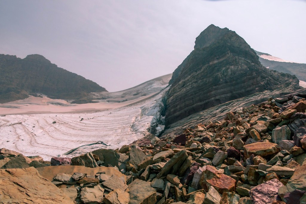 Sperry Glacier fills the left half of the photo, with a large rock prominence filling the right side. The glacier appears to be wrapped around the rock prominence. 