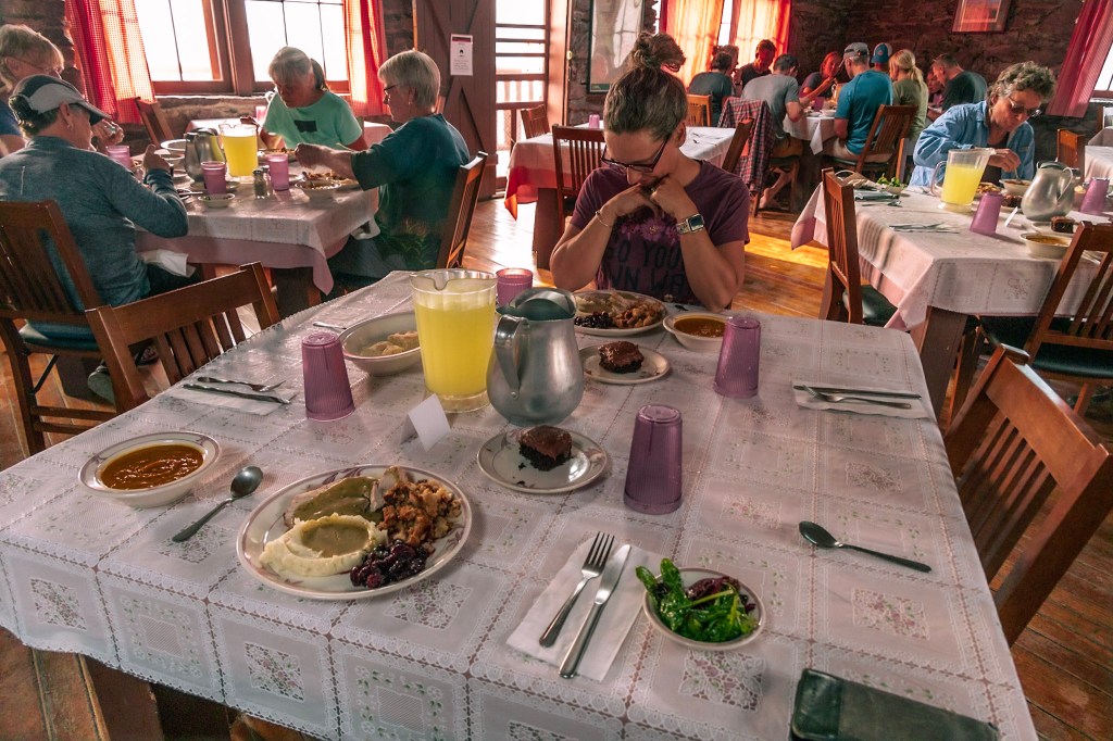 A dining table at Sperry Chalet dining hall set with a very large meal fills the frame. Laura sits on the far side of the table, looking lovingly down at her food. Other diners fill the background. 