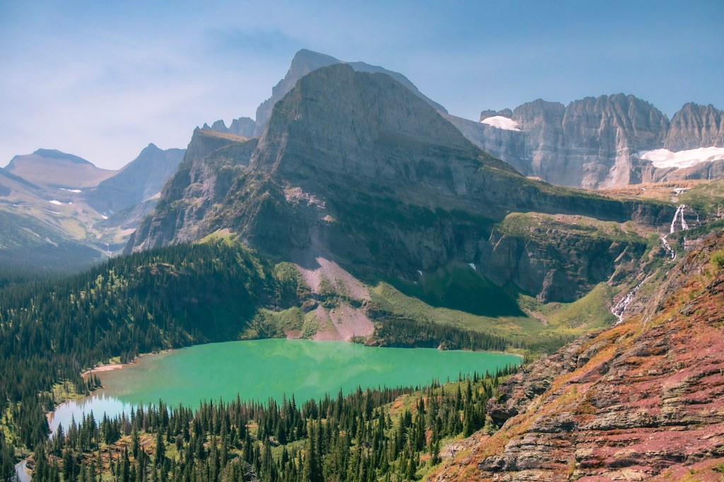 A bright teal glacial lake sits at the bottom of a valley. A waterfall cascades into the lake down hundreds of vertical feet of rock on the right side of the frame. A half-dome-shaped rock face looms over the scene at the center of the frame. Hazy blue sky is visible above. 