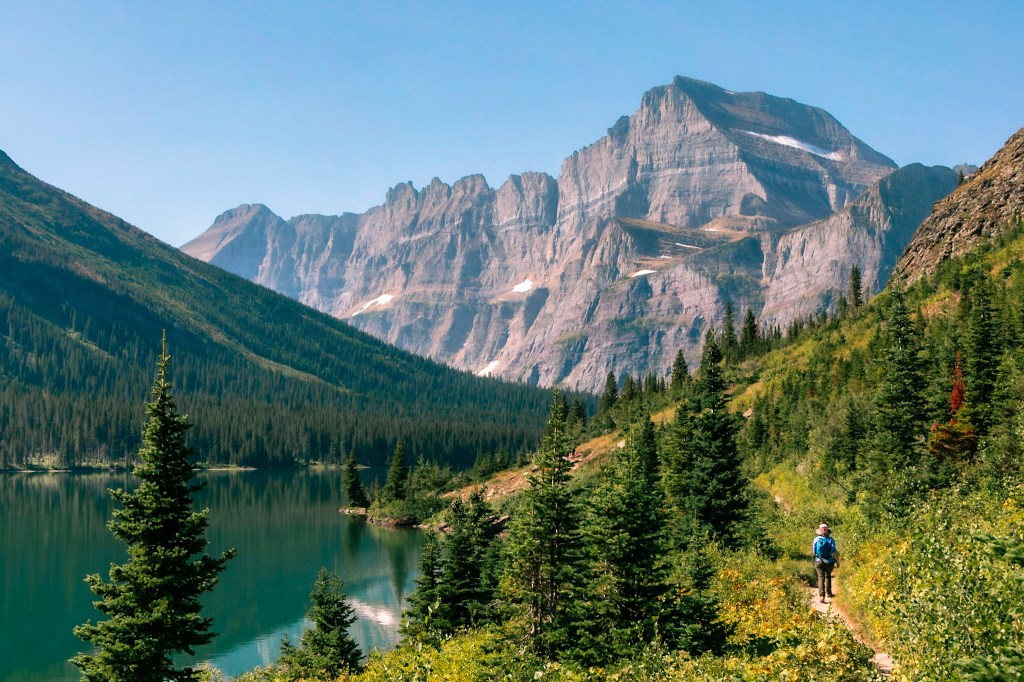 Laura stands on a trail in the bottom right of the frame, with the teal waters of a glacial lake to her left. A sheer rock mountainside rises above them, stretching into a hazy blue sky. 