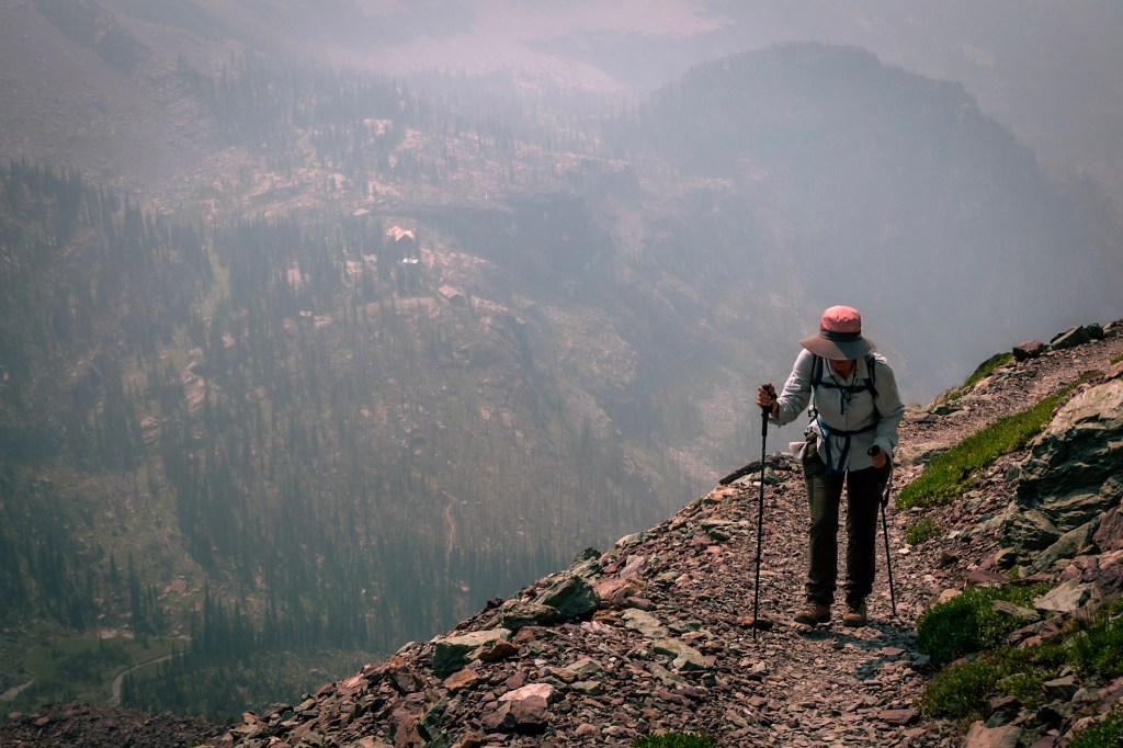 Laura walks toward the camera on the right side of the photo, holding hiking poles and looking tired. She stands on a rocky path that appears to drop away steeply just to the left. Obscured by haze, Sperry Chalet can be seen below and at a great distance.
