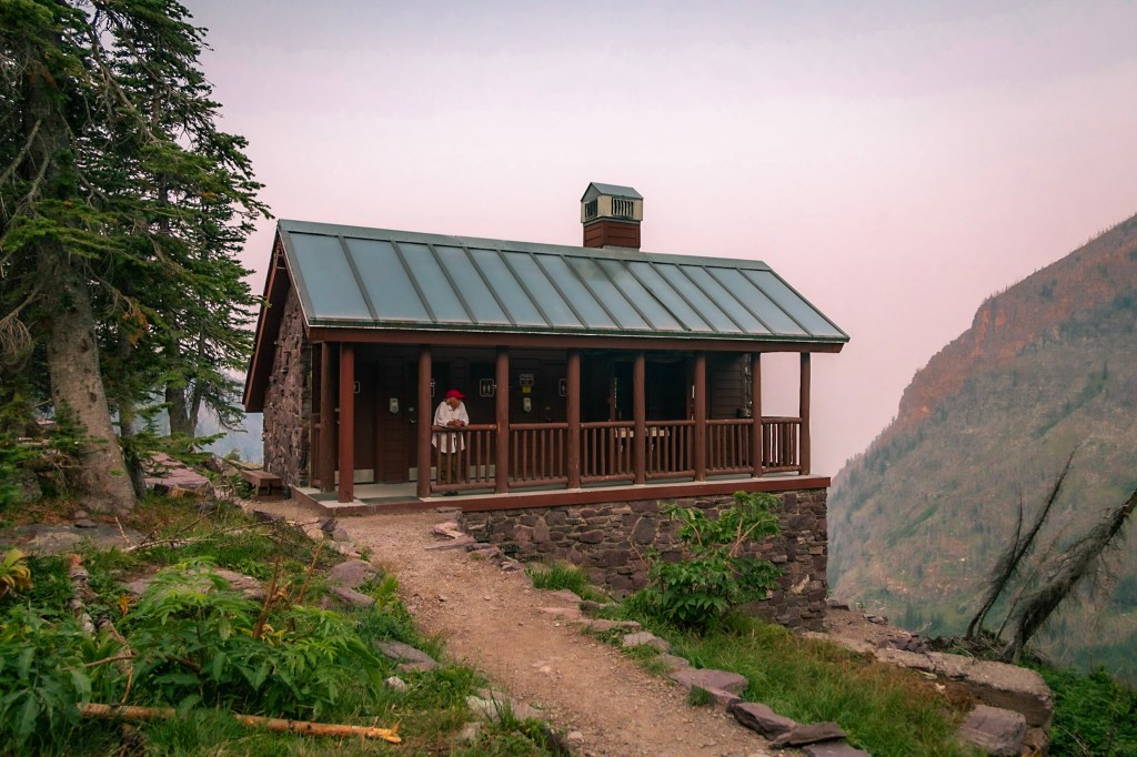 A stone building with a steeply sloped roof sits on the edge of a steep hill at the center of the photo. The building has a front porch with a railing, behind which can be seen four doors and two sinks. This is the Sperry Chalet pit toilet building.