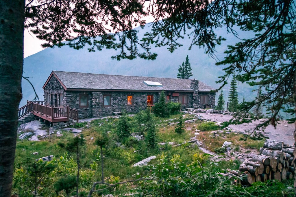 A long stone building stretches across the center of the photo with orange light glowing out of several windows. The building sits on the edge of a cliff, which drops off behind it. The photo is framed by the trunk and branches of a fir tree. 