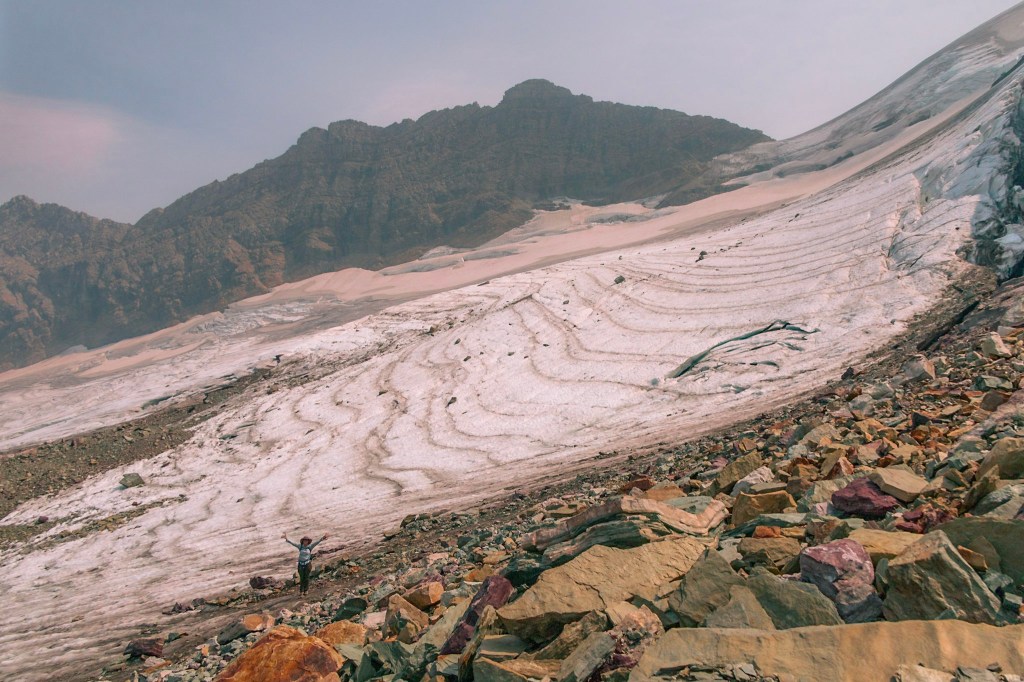 Sperry Glacier stretches from the top left to bottom right of the photo with colorful rocks in the foreground. Laura can be seen, very small, at the bottom left of the photo, on the edge of the glacier, with her arms raised victoriously.