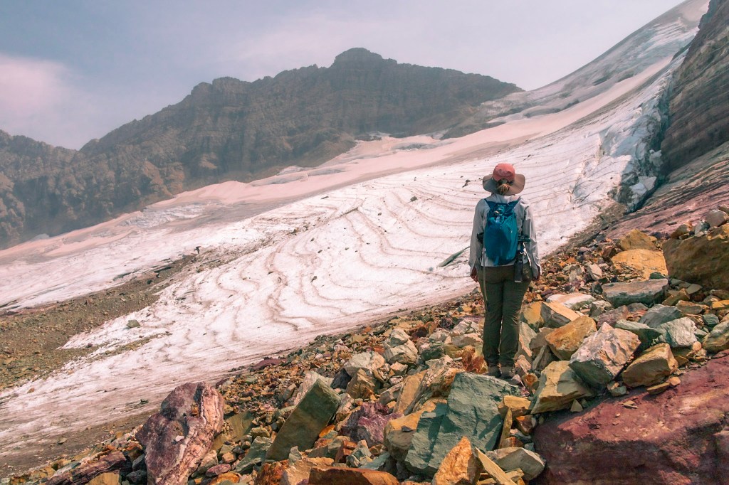 Laura stands on colorful rocks in the center right of the photo, back to the camera, looking toward Sperry Glacier which stretches diagonally from the top right to bottom left of the photo. 