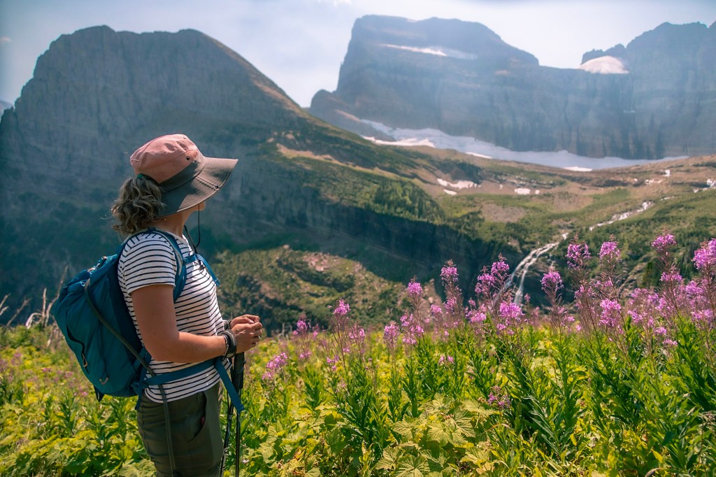 Laura stands on the left side of the frame, with brilliant magenta fireweed flowers blooming to the right. In the background, a waterfall cascading down rocky mountains is just visible. 