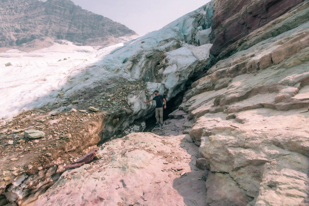 A similar image of Dustin standing in the gap between Sperry Glacier at the rock, touching the ice.
