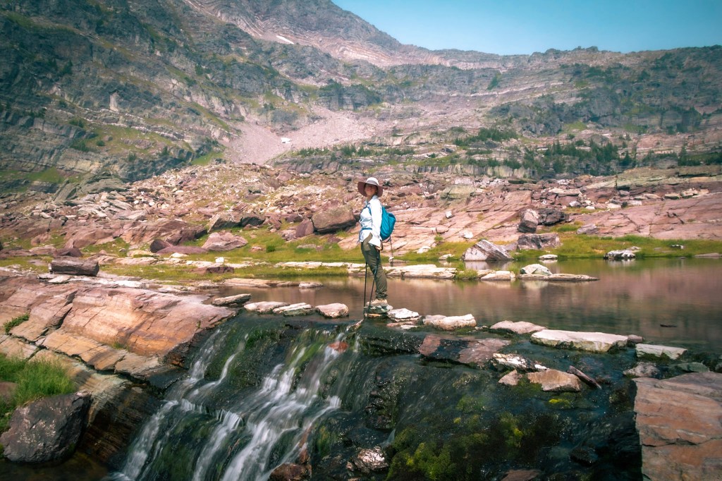 Laura stands at a distance in the center of the frame, looking over her shoulder at the camera. She stands on rocks crossing the edge of Feather Woman Lake, which turns into a waterfall just below her feet. Mountains rise in the background. 