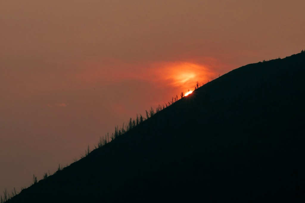 The sun sets behind the slope of a black mountain at the center of the frame. Thick smoke causes the sun to appear bright orange. They sky is very dark gray.