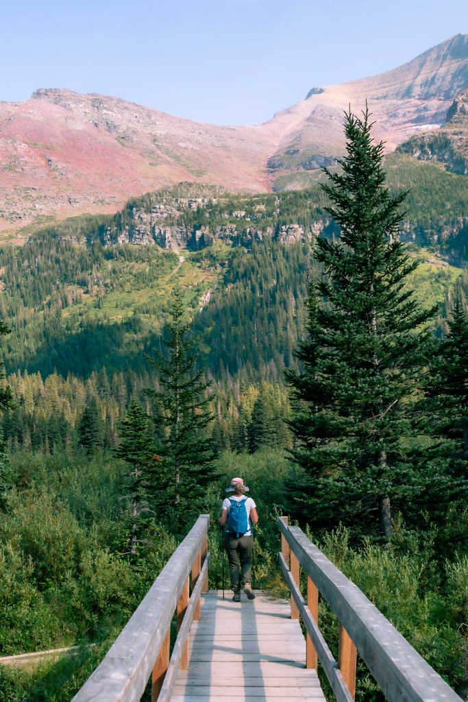 Laura stands facing away from the camera in the bottom third of the image, framed by a wooden bridge that she's just reached the end of. A fir and spruce forest stretches out before her. The trees climb up the sides of a rocky valley, with red-and-grey bare rock mountain tops visible at the top of the image. 