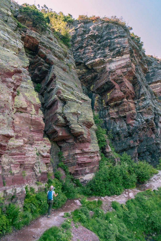 Laura walks along a rocky trail at the bottom of the image. A craggy cliff of red, green, and black rocks rises sharply above her. 