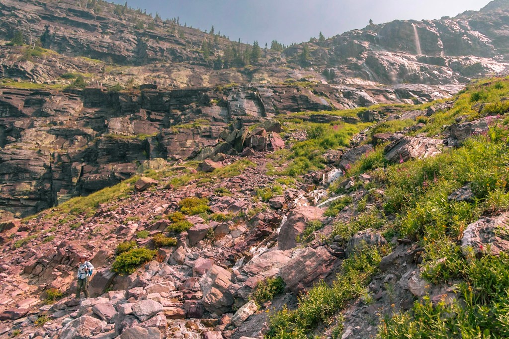 A rocky landscape with a waterfall running diagonally from top right to bottom center. Laura, far away, can be seen bottom left. 