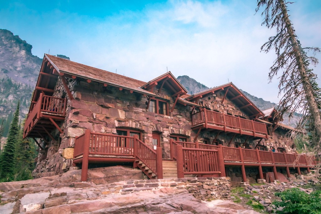The Sperry Chalet dorm building, a building with a peaked roof and gables fills the photo. A log porch lines the lower level with a smaller porch centered on the upper level. A mountain rises steeply behind it. 
