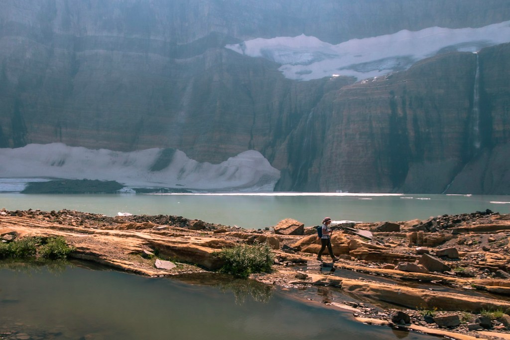 In the bottom half of the image, Laura walks along a row of rock that appears to be a peninsula or island in the middle of the glacial lake. In the background, the snow and ice of two glaciers are visible, with melting water cascading down a sheer vertical rock face into the lake. 