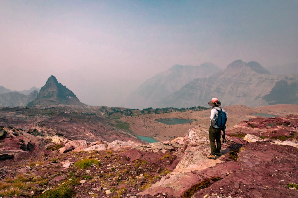 Laura stands bottom right, back to camera, looking out at the edge of the rock shelf that holds Sperry Glacier. None of the glacier can be seen, only several small pools in the background of melted glacier. The far background is thick with smoke.