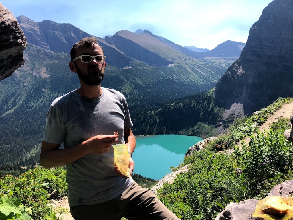 Dustin stands in the middle of the frame, holding a yellow bag of snacks. His face is obscured by shadow, but you can see some damp marks on his t-shirt. A bright teal glacial lake and some tree-covered mountains are visible in the background. 