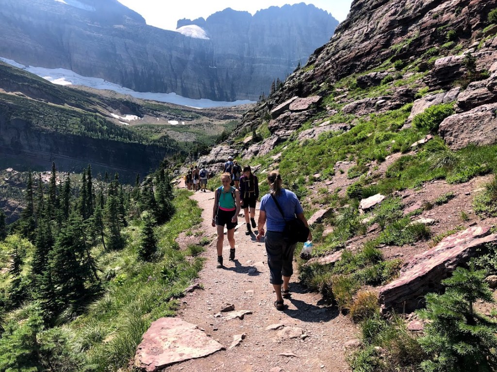 A line of about a dozen people pass each other on a mountain trail in the middle of the frame. A rocky slope is to the right, while the hazy outline of a cliff rises above the scene in the background. 