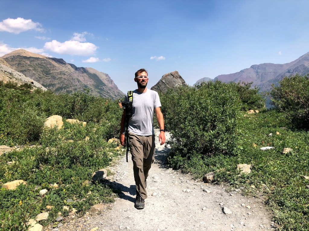 Dustin stands in the center of the image, striding along a rocky trail. Some green bushes fill the landscape on either side, with some bigger mountains in the background. 