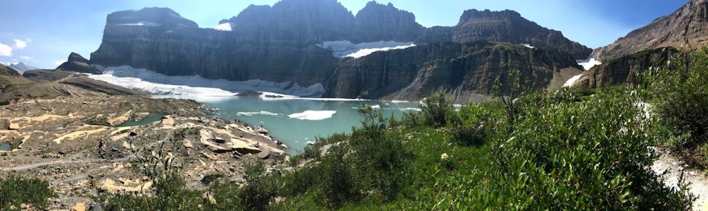 A panoramic view with a teal glacial lake, dotted with snow and ice, at the center of the image. A mass of rocky peaks rises above it. Some green undergrowth and a bare rocky plateau are in the foreground. 