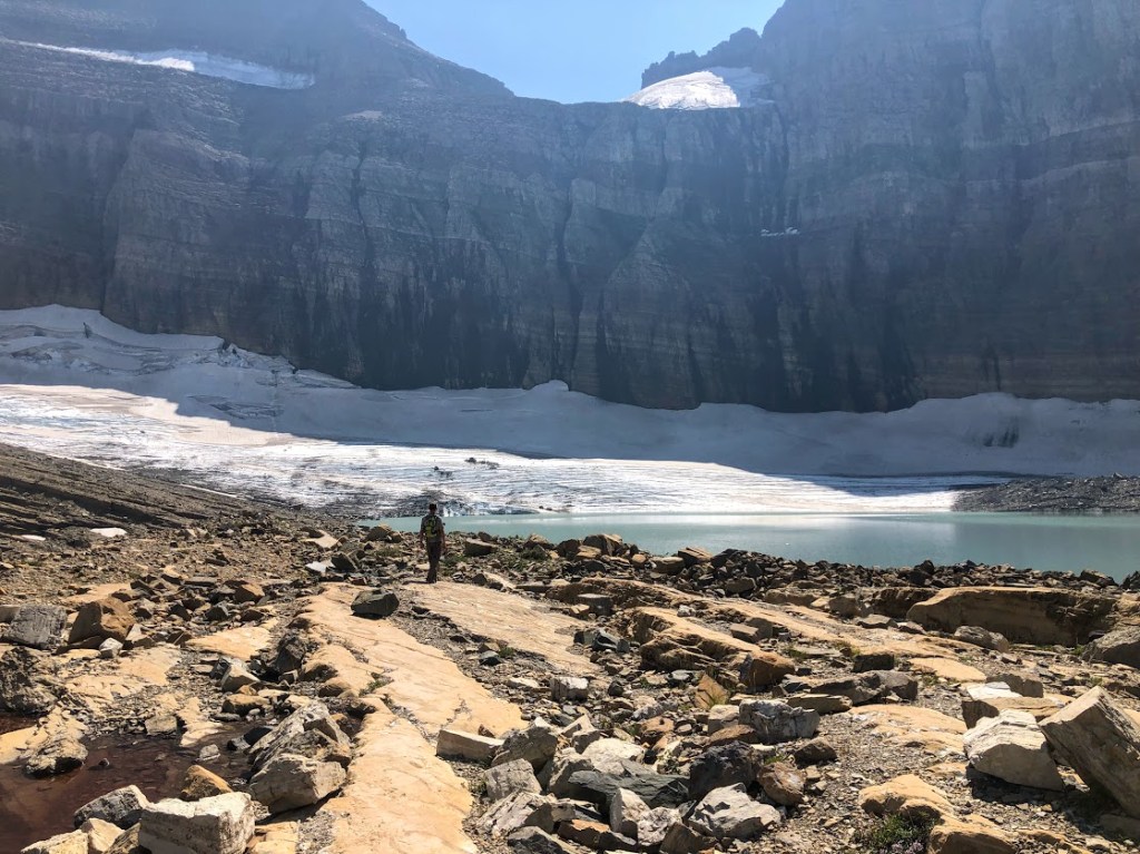 The rock wall of a cliff fills the background of the image, with a rocky plateau in the foreground. In between, you can make out a lake, a glacier, and someone walking toward them. 