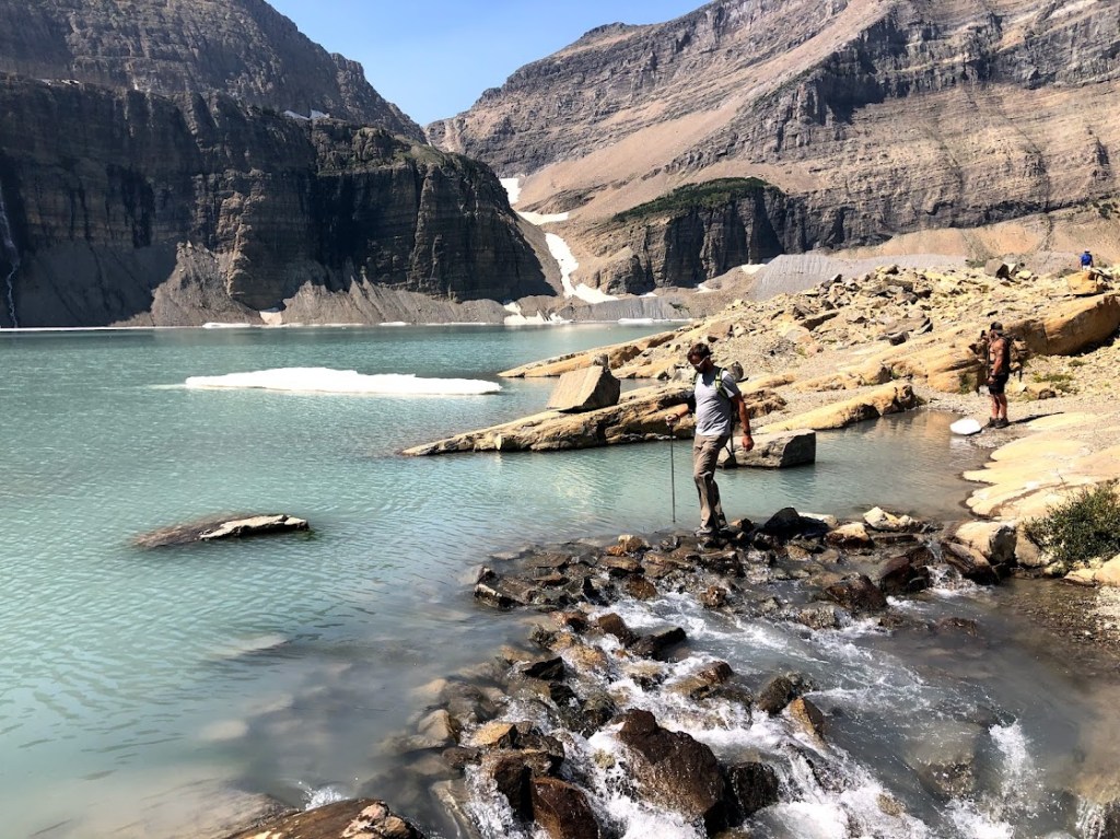 Dustin navigates the same rocks from the opposite view. The lake, dotted with snow and ice, is visible on the left. Rocky mountains rise above the scene in the background. The water from the lake cascades over the rocks and down to the right. 